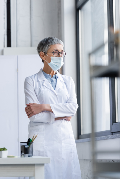 Beyaz önlüklü ve tıbbi maskeli olgun bir doktor bulanık önplanda pencere ve masanın yanında duruyor.  - Fotoğraf, Görsel