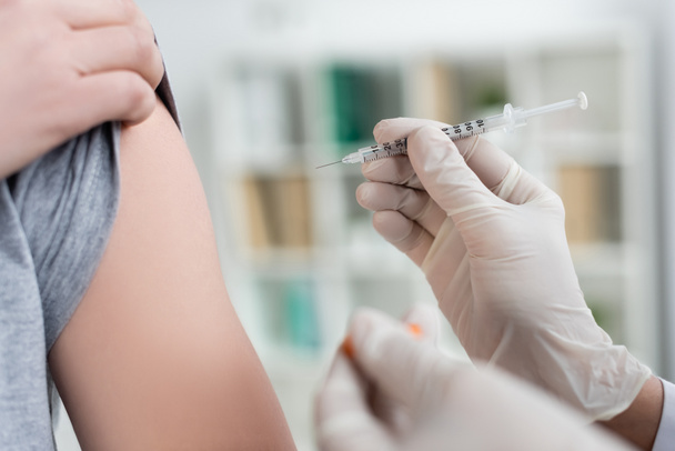 Oříznutý pohled na lékaře v latexových rukavicích držících injekční stříkačku a vakcínu na rozmazaném popředí poblíž pacienta  - Fotografie, Obrázek
