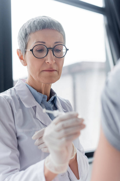 Mature doctor holding syringe on blurred foreground near patient  - Photo, Image