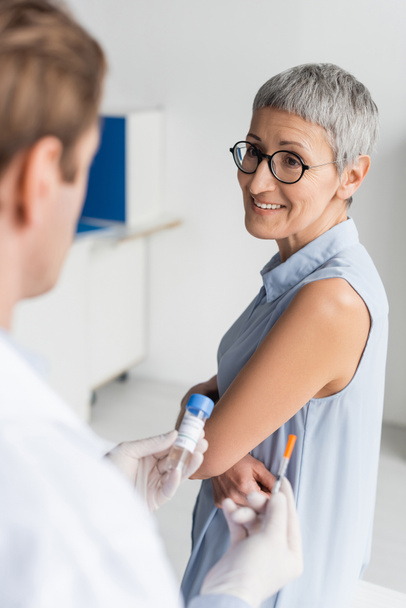 Mulher sorridente de pé perto do médico com vacina e seringa em primeiro plano turvo  - Foto, Imagem