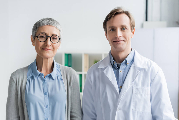 volwassen vrouw en arts in witte jas kijken naar camera in kliniek  - Foto, afbeelding