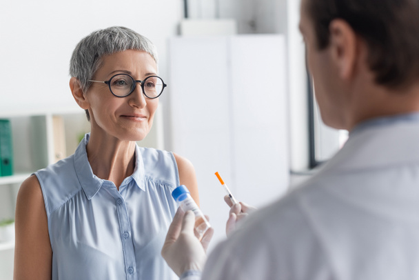 Glimlachende vrouw die naar een arts kijkt met spuit en vaccin op wazige voorgrond  - Foto, afbeelding