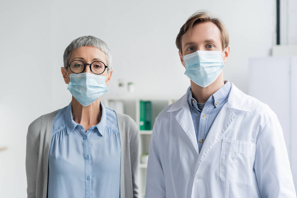 Mujer madura y médico en máscaras médicas mirando a la cámara en la clínica  - Foto, Imagen