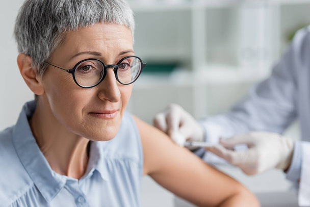 Mature woman sitting near doctor during vaccination on blurred background  - Φωτογραφία, εικόνα