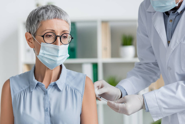 Mujer madura en máscara médica y médico haciendo la vacunación en la clínica  - Foto, Imagen