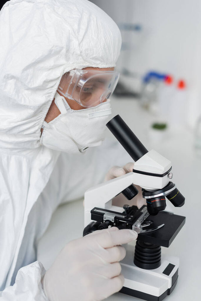 Ученый в латексных перчатках и очках с помощью микроскопа в лаборатории  - Фото, изображение