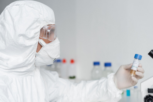 Wetenschapper in beschermende kleding en brillen met vaccin op wazige achtergrond  - Foto, afbeelding