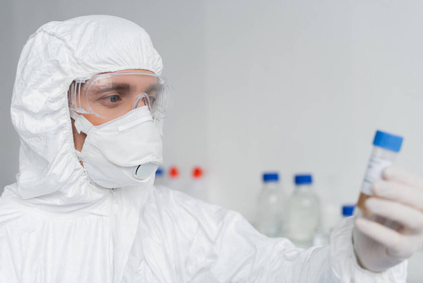 Wissenschaftler in Schutzanzug und Maske mit Impfstoff im verschwommenen Vordergrund  - Foto, Bild