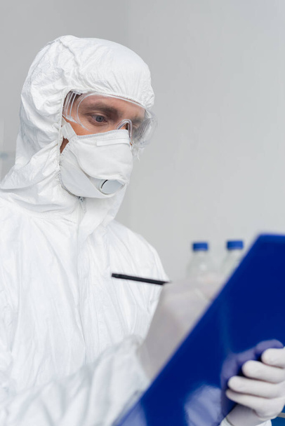 Científico en traje de materiales peligrosos y la escritura máscara médica en el portapapeles en primer plano borrosa  - Foto, Imagen