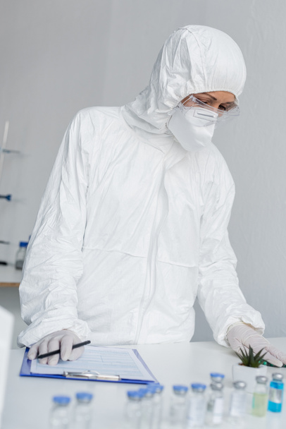 ワクチンの近くに立つハズマットスーツの科学者と研究室でぼやけた前景にクリップボード  - 写真・画像