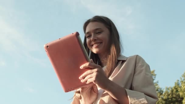 Lage hoek POV van jonge vrolijke blanke vrouw glimlachend op de voorgrond van groene bomen en blauwe lucht buiten, met behulp van tablet computer in roze geval op zonnige dag, dan kijken op camera - Video
