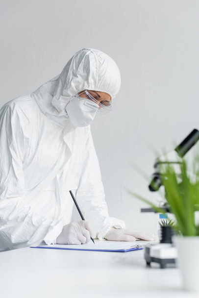 Ученый в защитном костюме пишет на планшете возле микроскопа на размытом переднем плане  - Фото, изображение
