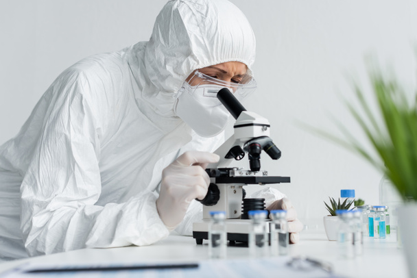 Wetenschapper in hazmat pak werken met microscoop in de buurt van vaccins op wazige voorgrond  - Foto, afbeelding