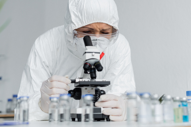 Scientist in goggles and hazmat suit looking at microscope near vaccines on blurred foreground  - Photo, Image