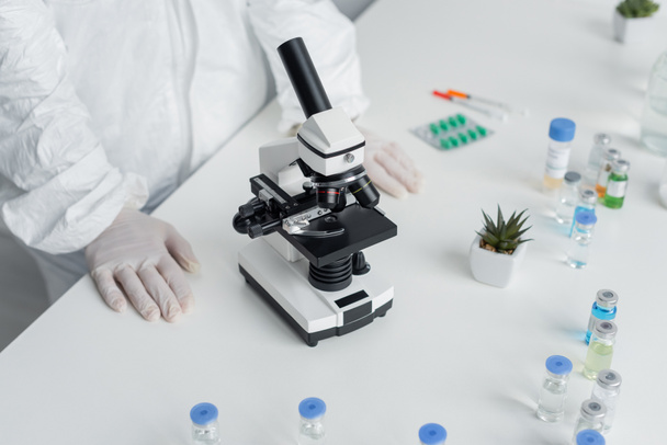 Vista ritagliata dello scienziato in tuta Hazmat in piedi vicino al microscopio e vaccini sul tavolo  - Foto, immagini