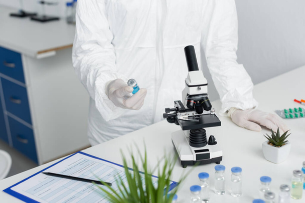 Vista recortada del científico en traje de materiales peligrosos que sostiene la vacuna cerca del microscopio y el portapapeles  - Foto, Imagen