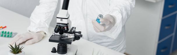 Vista recortada del científico en uniforme protector que sostiene la vacuna cerca del microscopio y las píldoras, bandera  - Foto, imagen