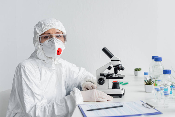 Wetenschapper in beschermend masker en bril kijkend naar camera nabij microscoop, vaccins en klembord - Foto, afbeelding