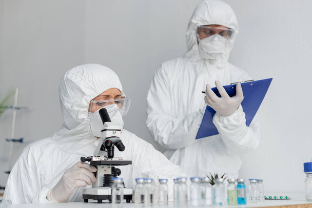 Científico en uniforme protector usando microscopio cerca de colega y vacunas en primer plano borroso  - Foto, Imagen
