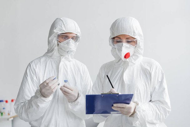 Científicos en trajes de materiales peligrosos que trabajan con vacunas y portapapeles en laboratorio  - Foto, Imagen
