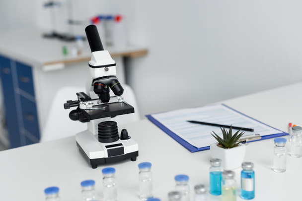 Microscope near vaccines and clipboard on blurred background in laboratory  - Φωτογραφία, εικόνα