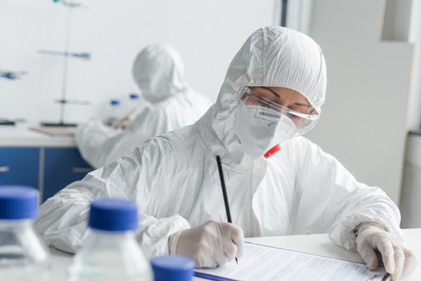 Scientifique écrivant sur presse-papiers près d'un collègue sur fond flou en laboratoire  - Photo, image