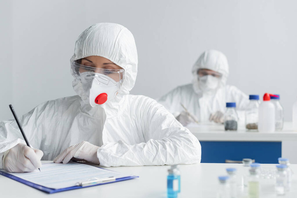 Scientist in protective suit and latex gloves writing on clipboard near vaccines and colleague  - Photo, Image