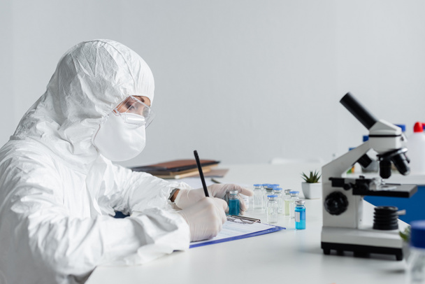 Scientist in protective uniform writing on clipboard near microscope and vaccines  - Photo, Image