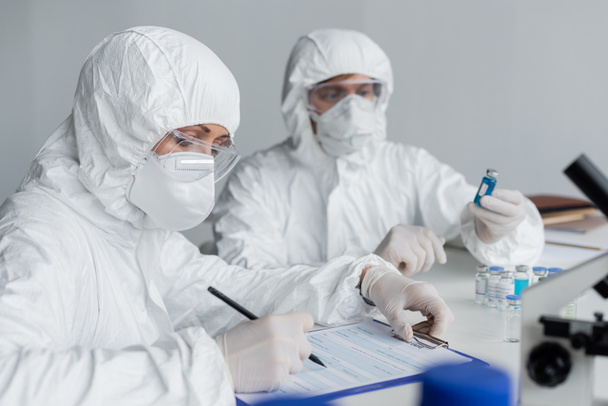 Wissenschaftler schreibt auf Klemmbrett neben Kollege mit Impfstoffen auf verschwommenem Hintergrund  - Foto, Bild
