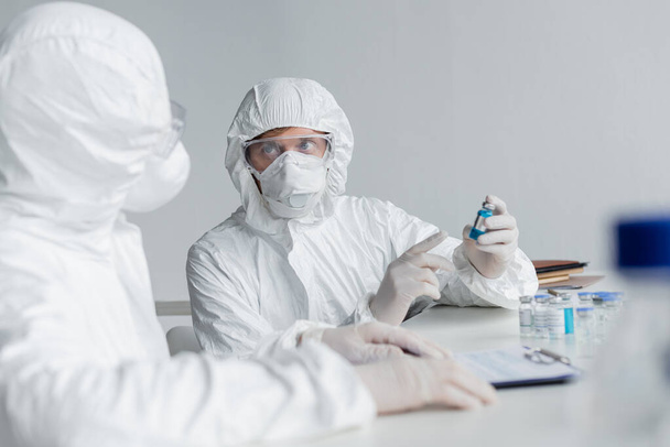 Koruyucu giysili bilim adamı, laboratuardaki bulanık ön planda iş arkadaşının yakınındaki aşıyı işaret ediyor.  - Fotoğraf, Görsel