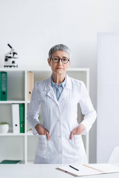 Mature doctor with hands in pockets of white coat looking at camera  - Photo, Image