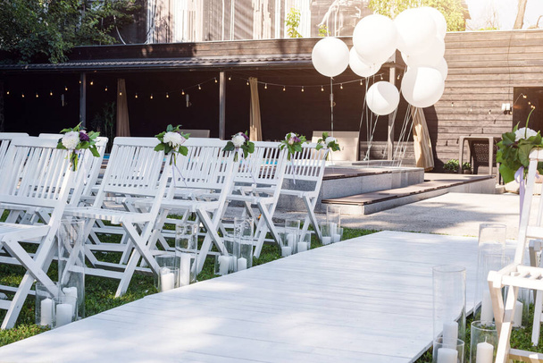 Ωραίος γάμος κανονίστηκε. Γαμήλια τελετή στον κήπο. Ξύλινες λευκές καρέκλες διακοσμημένες με λουλούδια, μπαλόνια και κεριά σε σειρές - Φωτογραφία, εικόνα