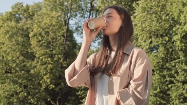 Низкоугольная талия молодой счастливой белой женщины, улыбающейся, сидящей в солнечном летнем парке, пьющей напиток из бумажной чашки, машущей рукой невидимому знакомому и уходящей - Кадры, видео