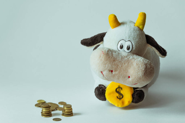 Ένα παιχνίδι με τη μορφή ενός χαμογελαστού ταύρου κρατά ένα κέρμα με ένα σύμβολο δολαρίου δίπλα σε χρυσά νομίσματα. - Φωτογραφία, εικόνα