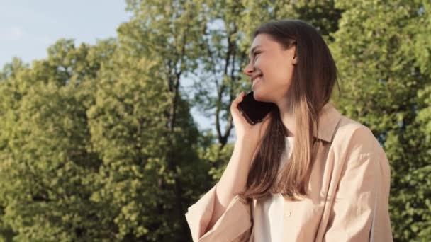 Tour de taille de jeune femme caucasienne heureuse souriante, assise dans un parc d'été ensoleillé, parlant sur un téléphone portable - Séquence, vidéo