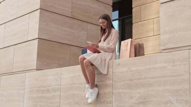 Μακρύ πλάνο της νεαρής Καυκάσιας γυναίκας φορώντας καλοκαιρινό φόρεμα και λευκά παπούτσια, κάθεται στη βεράντα του μεγάλου κτιρίου, χρησιμοποιώντας πιστωτική κάρτα, κάνοντας online πληρωμή μέσω του υπολογιστή tablet - Πλάνα, βίντεο