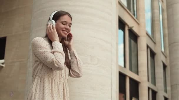 Low angle medium arc of long-haired cheerful Caucasian woman wearing summer dress and wireless over-ear headphones, smiling, enjoying music, moving along - Footage, Video