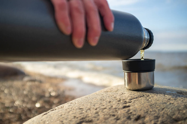 Hand pours a hot drink from a thermos into the lid of a cup on a stone on a sandy river bank on a spring day. - Photo, Image