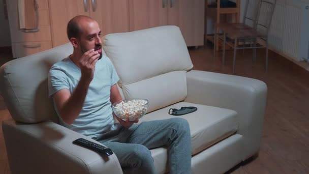 Kaukaski mężczyzna siedzi na kanapie z popcornem w ręku patrząc na serię filmów - Materiał filmowy, wideo