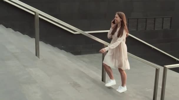 Brede tracking van bruinharige opgewonden blanke vrouw glimlachend, het dragen van zomerjurk en witte tennisschoenen, gaan de trap op buiten moderne gebouwen, praten op mobiele telefoon - Video
