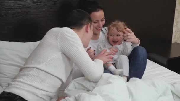 Young parents play with a small child. Happy family, child, daughter mom dad play paddle together in room on bed. Kid, mother hiding under covers, dad plays looking. Family laughing while playing game - Footage, Video