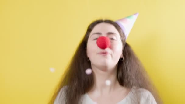 Retrato de una mujer alegre con una gorra de papel y una falsa nariz roja, que felizmente lanza confeti y aplaude. Fondo amarillo. Concepto del día tonto de abril - Metraje, vídeo