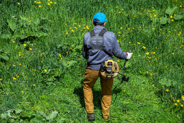 Работник газонокосилки стрижет зеленую траву летом при свете дня двухтактным триммером для двигателя в России. - Фото, изображение