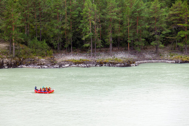Un team di persone che fanno rafting con giubbotti di salvataggio arancioni su un gommone di colori blu e gialli lungo un fiume di montagna sullo sfondo di una riva rocciosa con una foresta. - Foto, immagini