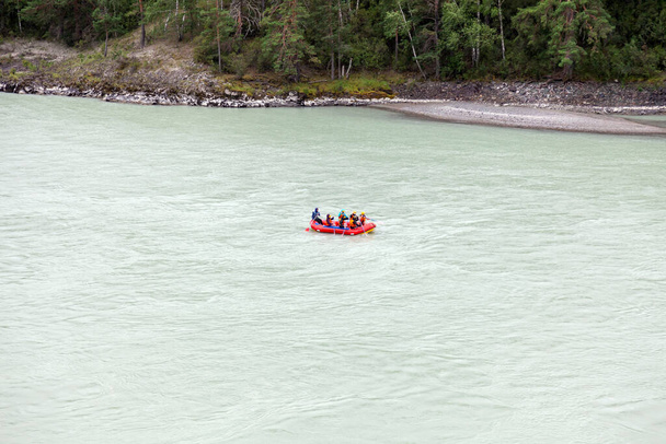 山の川沿いの青と黄色のゴムボートに乗って、森のある岩場の海岸を背景にオレンジのライフジャケットに身を包んだ人々のチーム. - 写真・画像