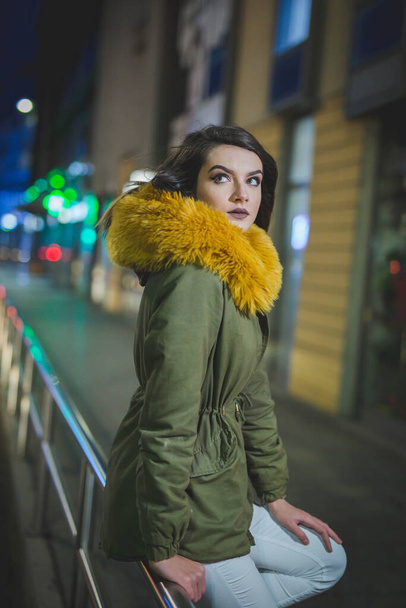 A vertical portrait of an attractive young female in a coat with yellow fur posing in a street at night - Фото, зображення