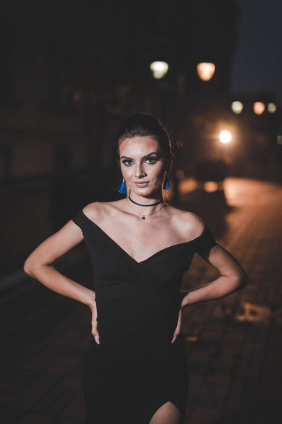 A vertical shot of an attractive young female in an elegant black dress posing in the street at night - Fotoğraf, Görsel