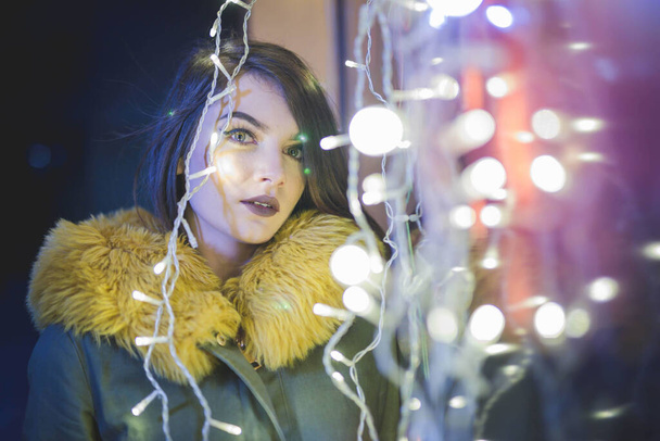 A portrait of an attractive young female in a coat with yellow fur posing near street lights at night - Fotoğraf, Görsel