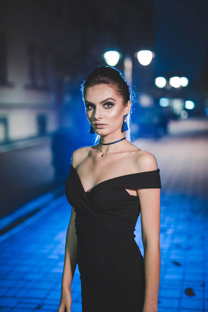 A vertical shot of an attractive young female in an elegant black dress posing in the street at night - Foto, Imagem