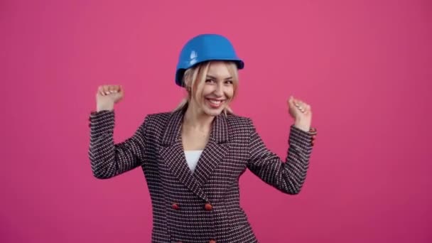 зрелая блондинка веселится и танцует с инженерами в шлеме на синем воротнике Молодая зрелая блондинка в розовой куртке. Изолированный на розовом фоне. - Кадры, видео